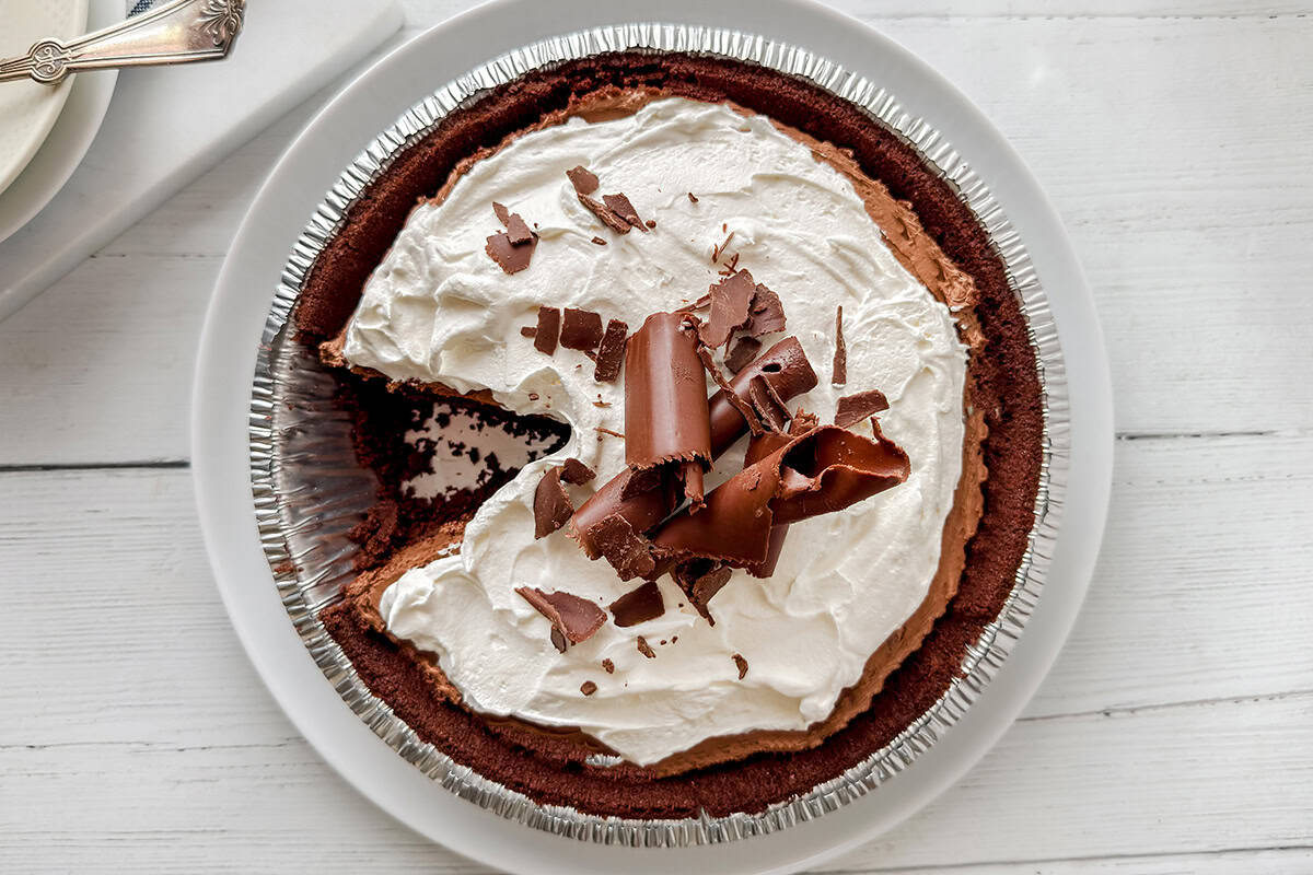 A chocolate pie with a slice missing, topped with whipped cream and chocolate shavings, sits on a white plate.