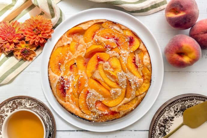 A peach cobbler cake dusted with powdered sugar sits on a white plate, surrounded by fresh peaches, orange flowers, a striped napkin, a cup of tea, and a plate with a yellow cake server on a white wooden table.