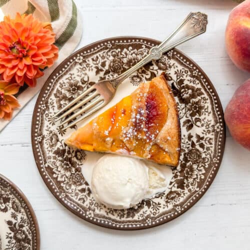 A plate with a slice of peach cobbler cake, a scoop of vanilla ice cream, and a fork. Beside the plate are two peaches, orange flowers, and a striped napkin on a white table.