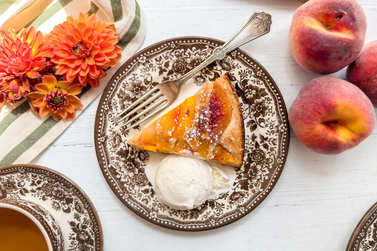 A plate with a slice of peach cobbler cake, a scoop of vanilla ice cream, and a fork. Beside the plate are two peaches, orange flowers, and a striped napkin on a white table.
