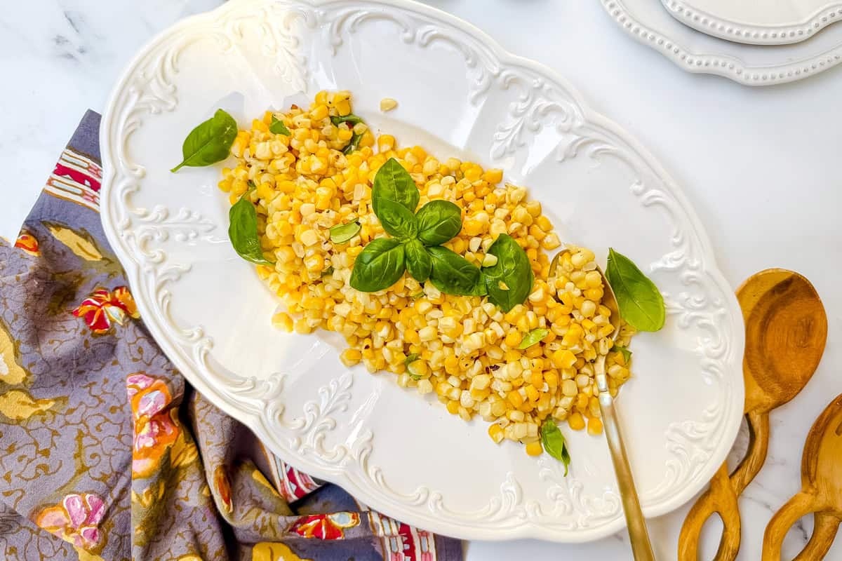 Sautéed Corn Recipe: Cooked corn kernels tossed in basil and butter, garnished with fresh basil leaves on a white oval platter, with a wooden spoon and patterned cloth nearby.