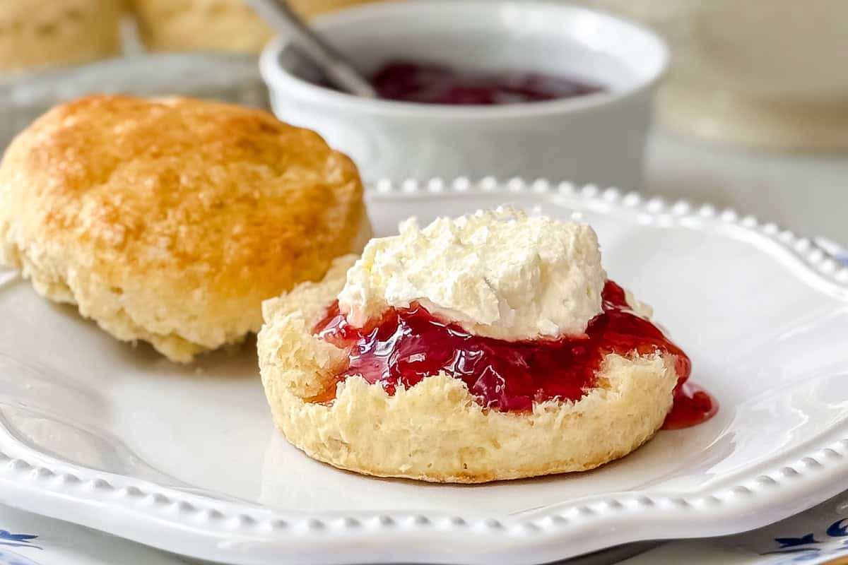 A scone cut in half on a white plate, topped with red jam and homemade clotted cream, with another whole scone and a bowl of jam in the background.