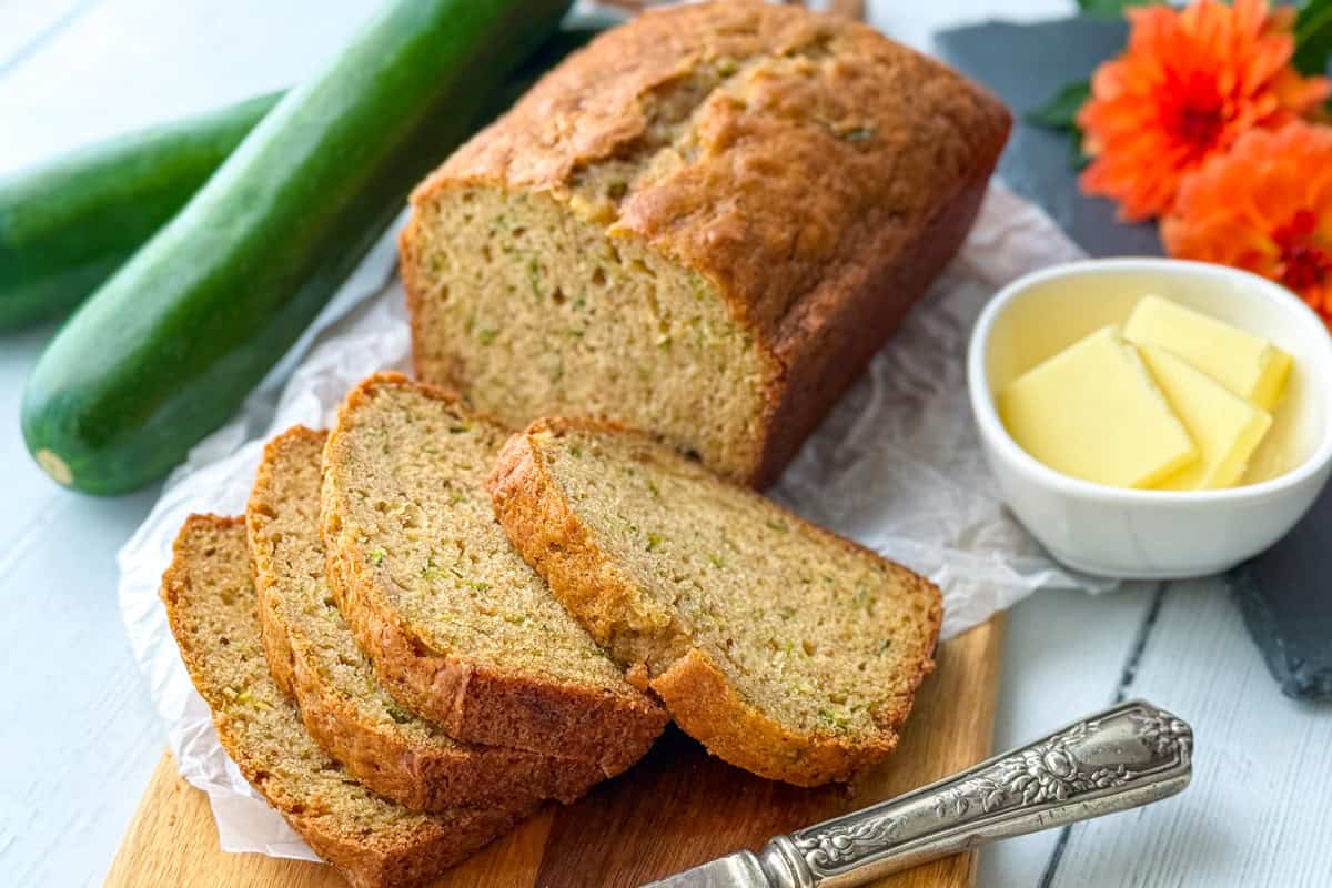 A single loaf of zucchini bread with several slices cut, placed on a wooden board beside a bowl of butter, a knife, zucchini, and orange flowers—an easy recipe perfect for any occasion.
