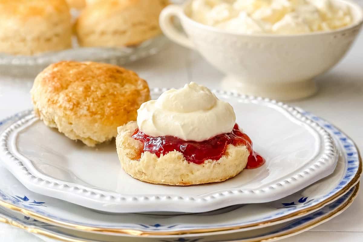 A scone topped with red jam and homemade mock clotted cream sits on a white plate, with another plain scone and a bowl of cream in the background.