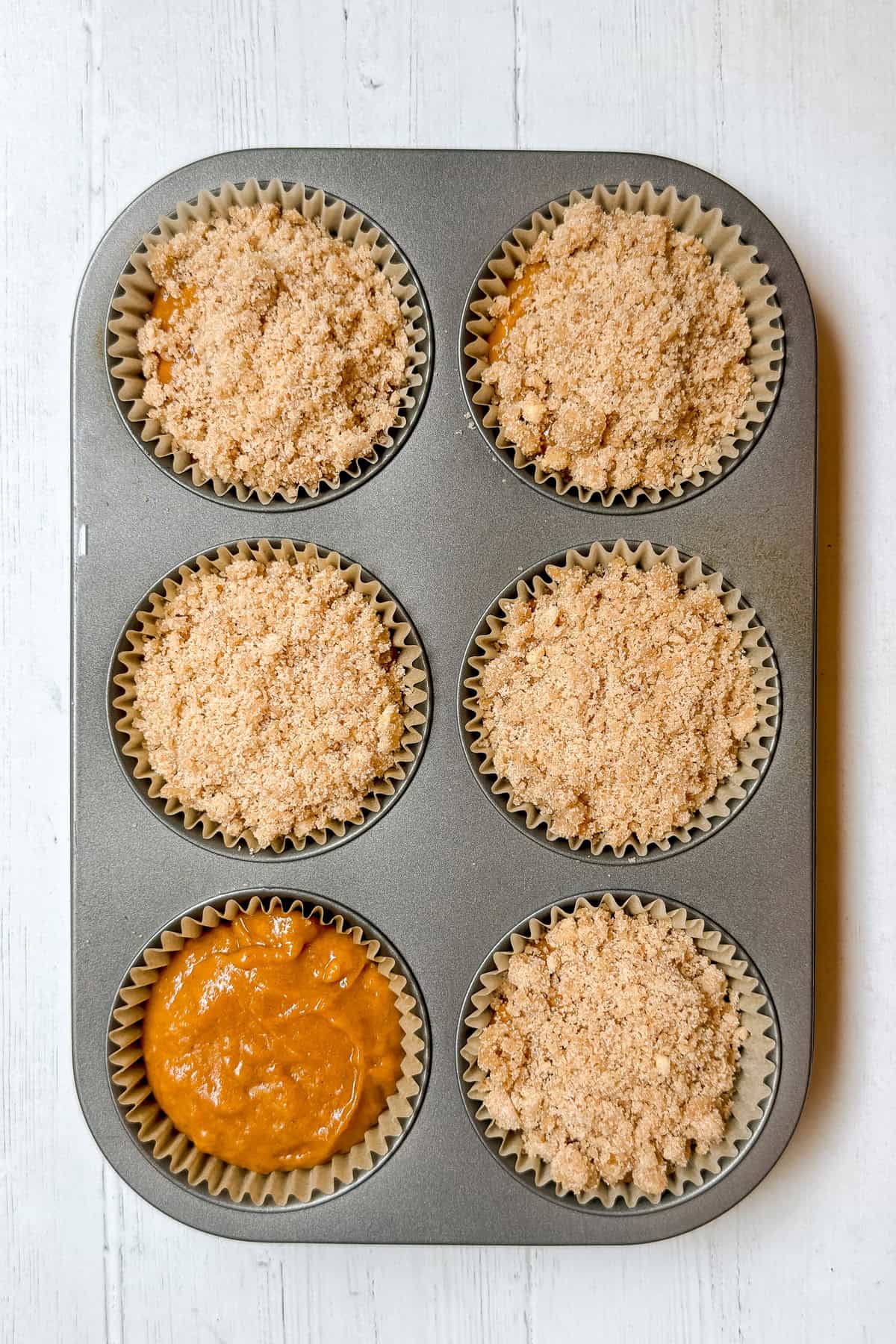 A muffin tin with six cups, five filled with batter topped with crumb topping and one with plain batter, sits on a white wooden surface, reminiscent of Panera's famous Pumpkin Muffins.
