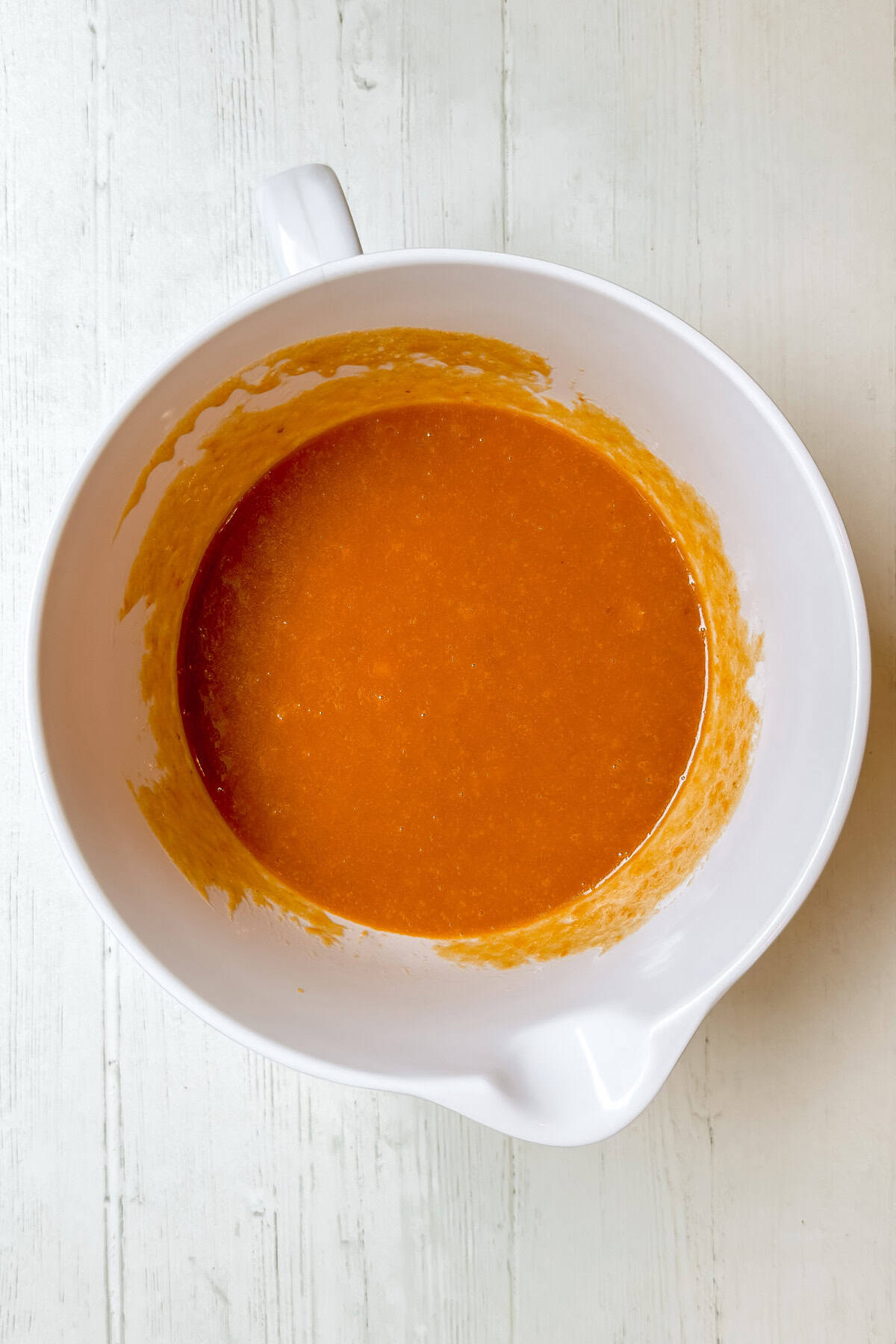 A white mixing bowl containing an orange liquid mixture, perfect for making Panera pumpkin muffins, sits on a light-colored wooden surface.