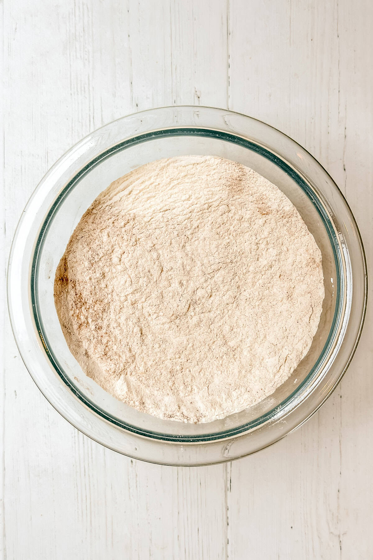 A clear glass bowl filled with a pale, finely ground powder, likely flour for Panera Pumpkin Muffins, sits on a white wooden surface.