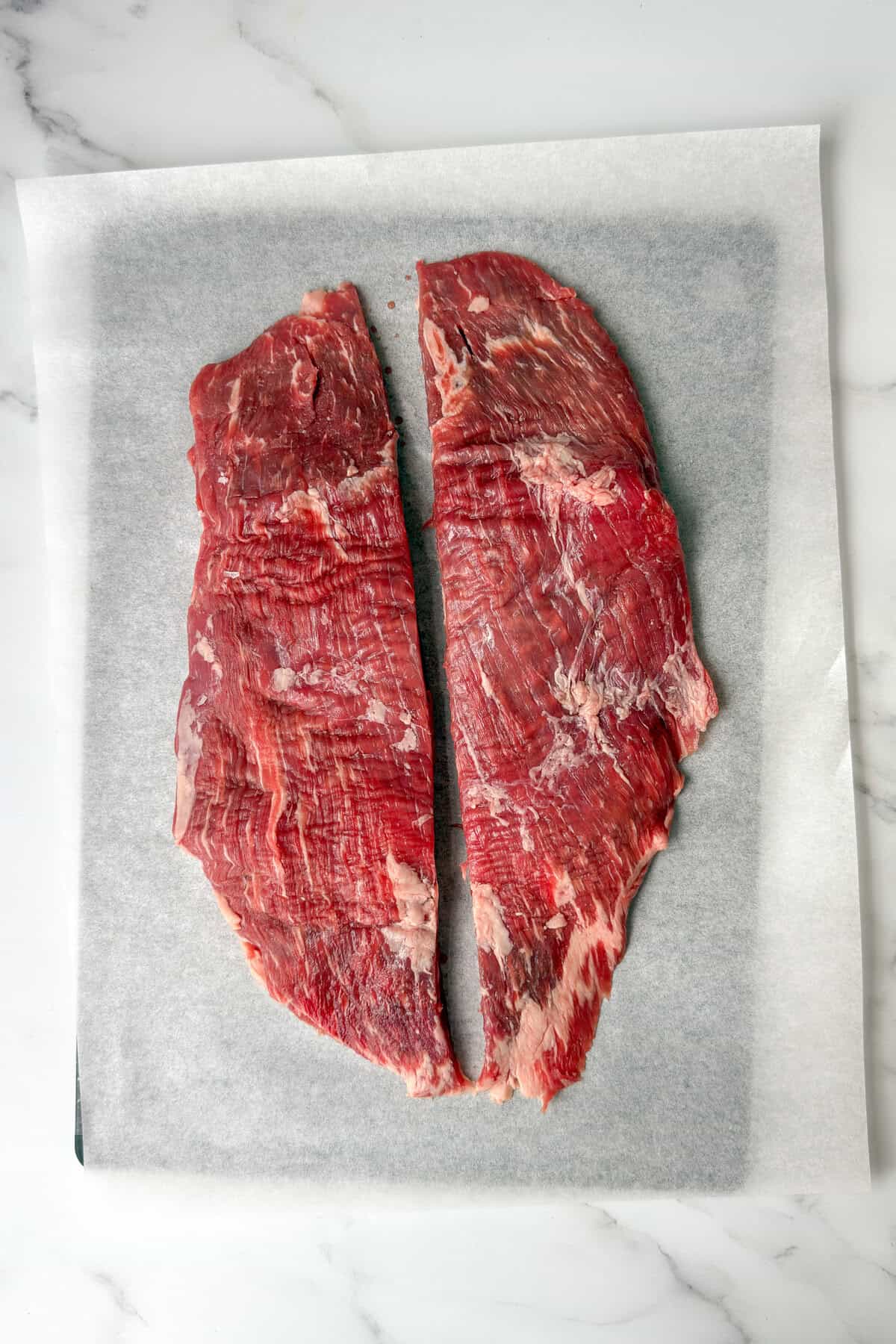 Two pieces of raw flank steak laid on parchment paper, ready for marinating.