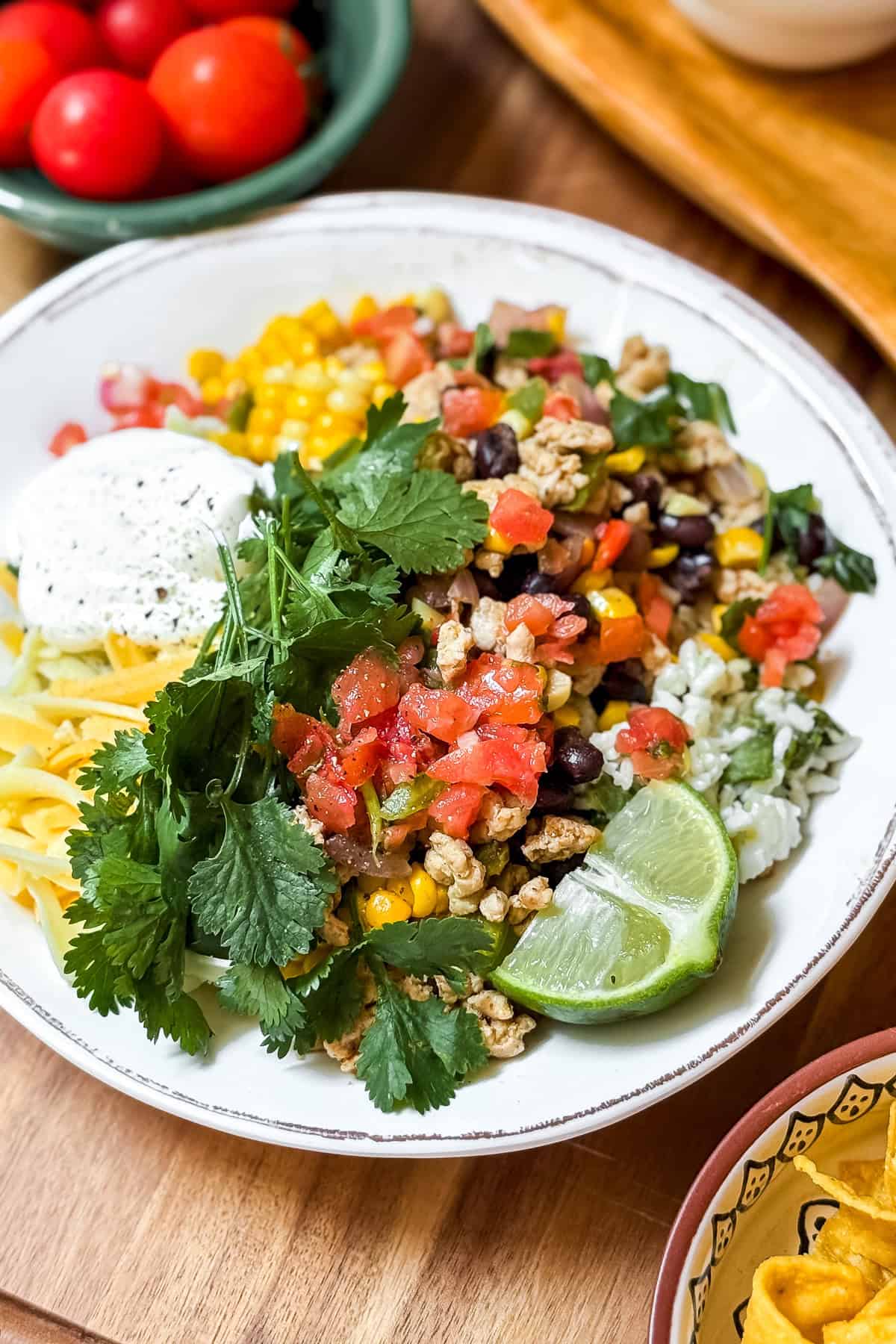 A plate with rice, black beans, corn, ground meat, shredded cheese, pico de gallo, sour cream, lime wedges, and fresh cilantro-perfect for Easy Chicken Taco Bowls. Cherry tomatoes are seen in the background.