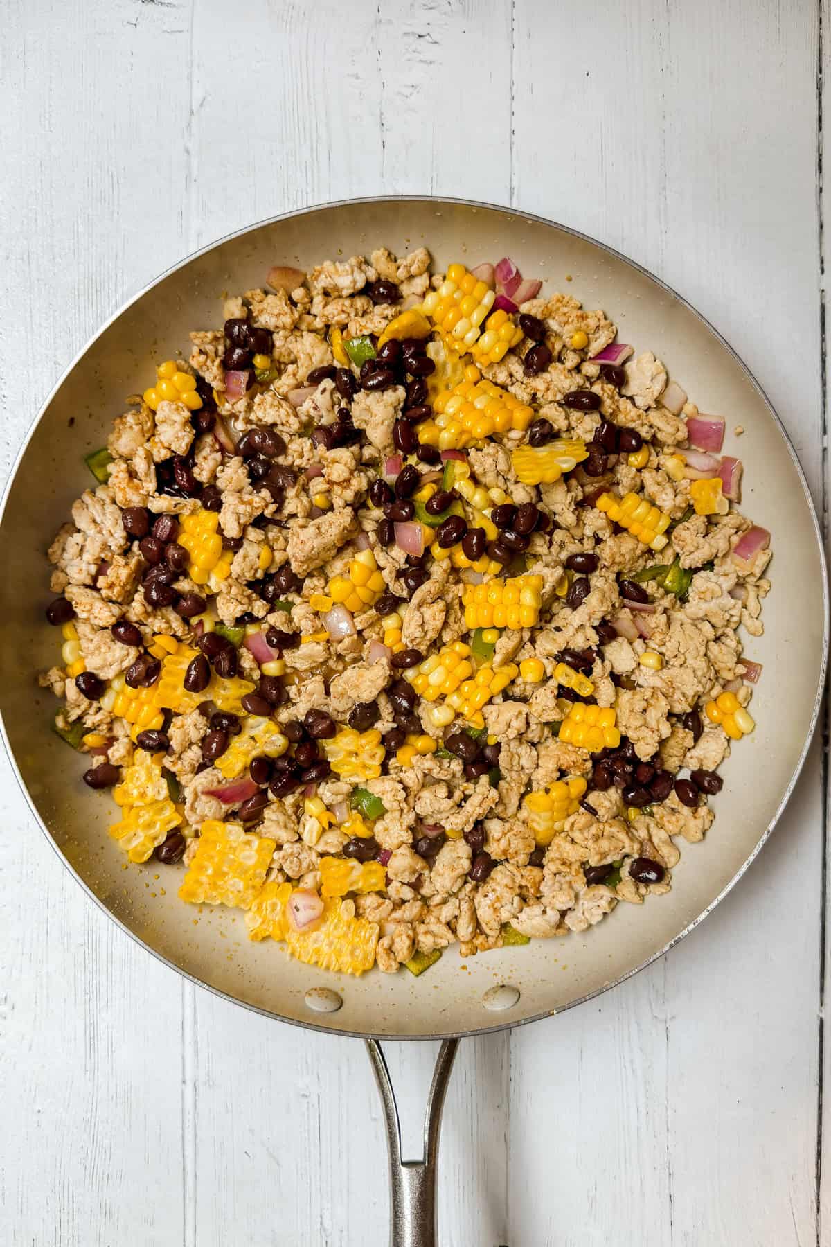 A skillet filled with cooked ground turkey, black beans, corn, chopped red onion, and green peppers-perfect for easy chicken taco bowls on a white wooden surface.