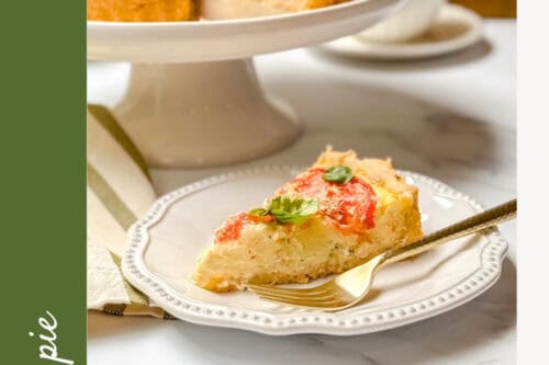 A slice of tomato basil pie with a garlic butter crust is served on a plate with a fork; the rest of the pie sits on a cake stand in the background.