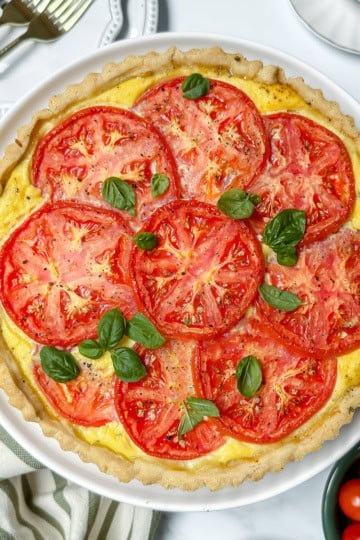 A baked tomato pie topped with fresh tomato slices and basil leaves, served in a white dish on a marble surface with a striped towel and utensils nearby.