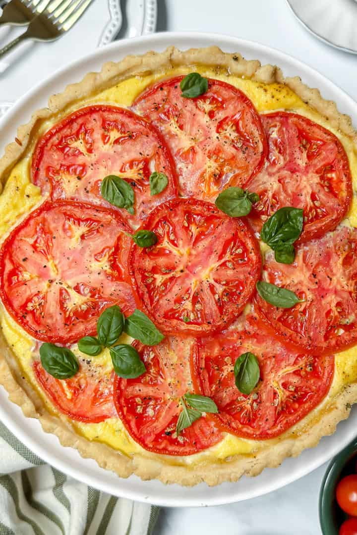 A baked tomato pie topped with fresh tomato slices and basil leaves, served in a white dish on a marble surface with a striped towel and utensils nearby.
