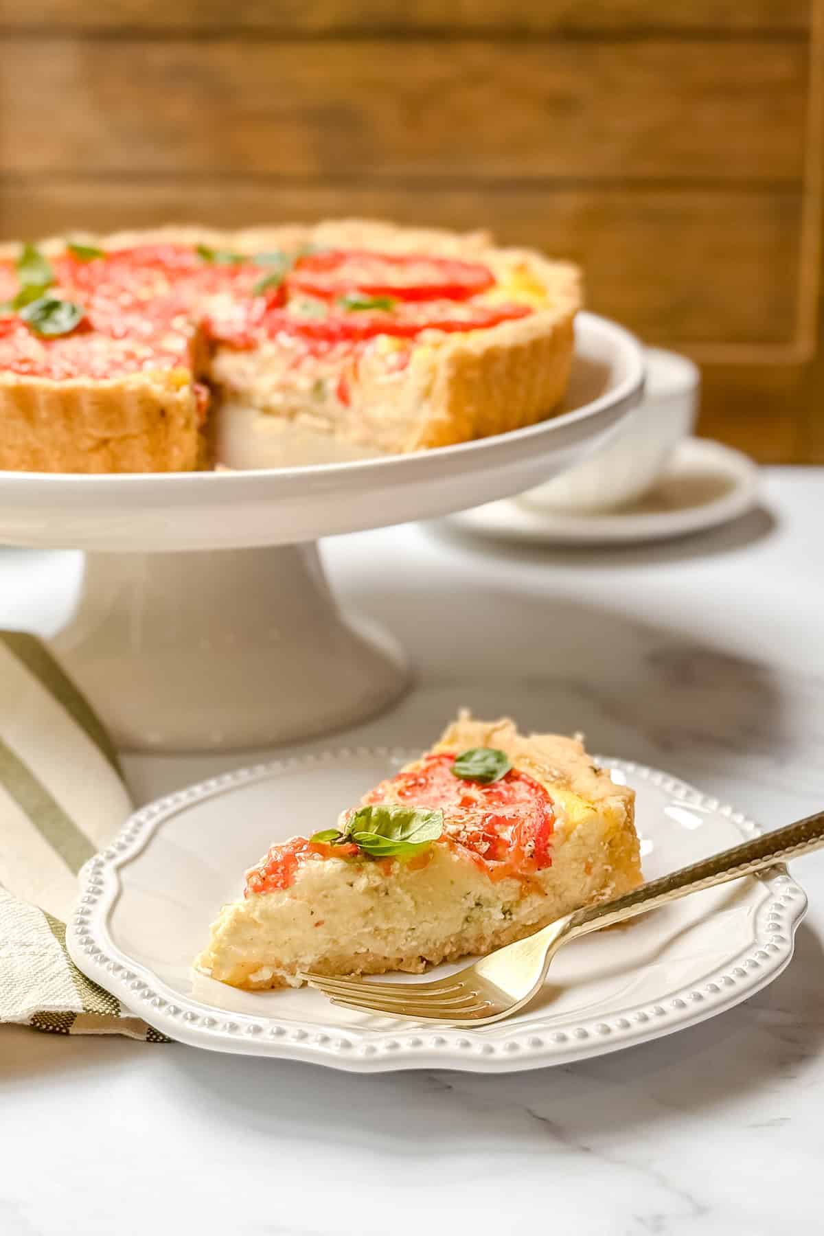 A slice of tomato basil pie-topped quiche sits on a white plate with a gold fork, while the rest of the quiche is on a cake stand in the background.