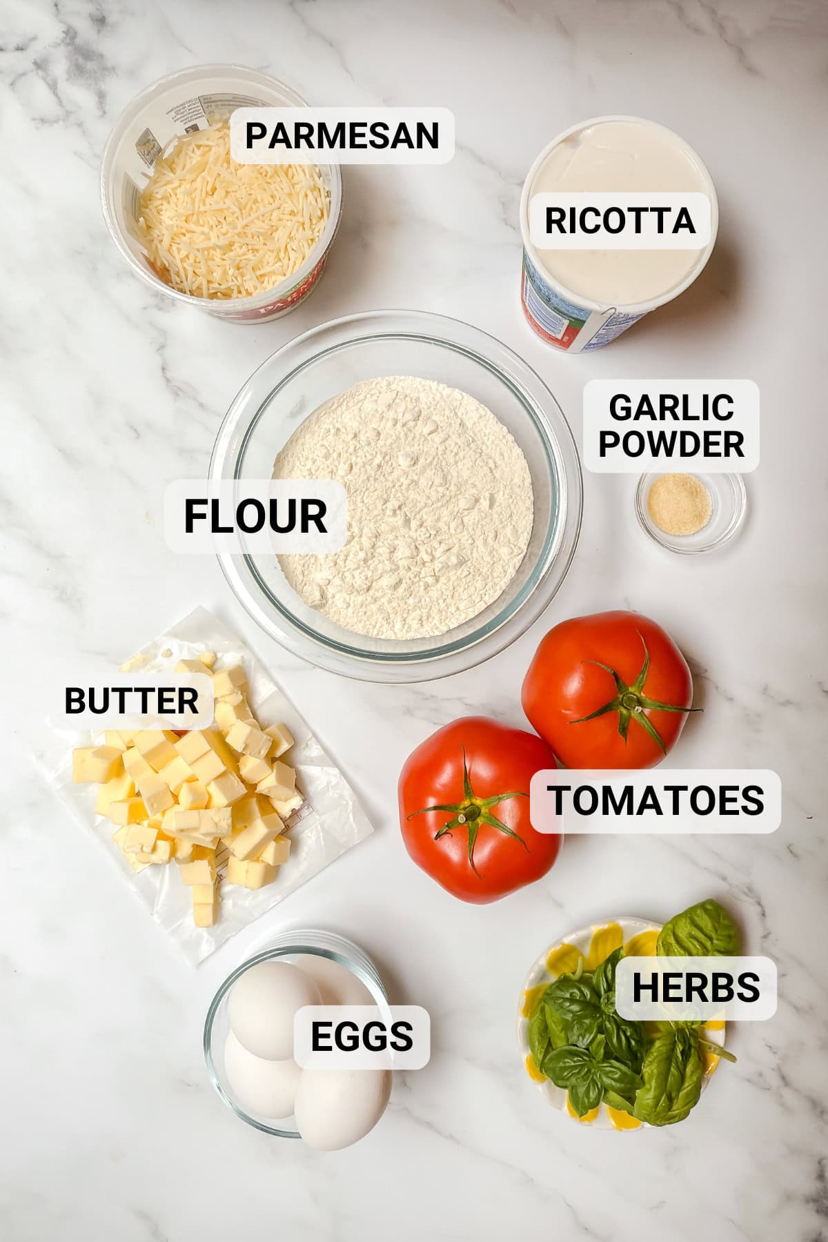 Top-down view of ingredients for a tomato basil pie on a marble surface: parmesan, ricotta, flour, garlic powder, butter cubes, eggs, tomatoes, and fresh herbs.