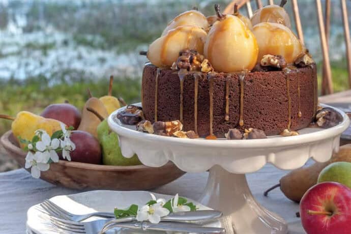 Chocolate cake topped with glazed pears and caramel drizzle on a white cake stand, surrounded by apples, pears, plates, and cutlery-perfect for an Autumn Afternoon Tea set outdoors by the water.