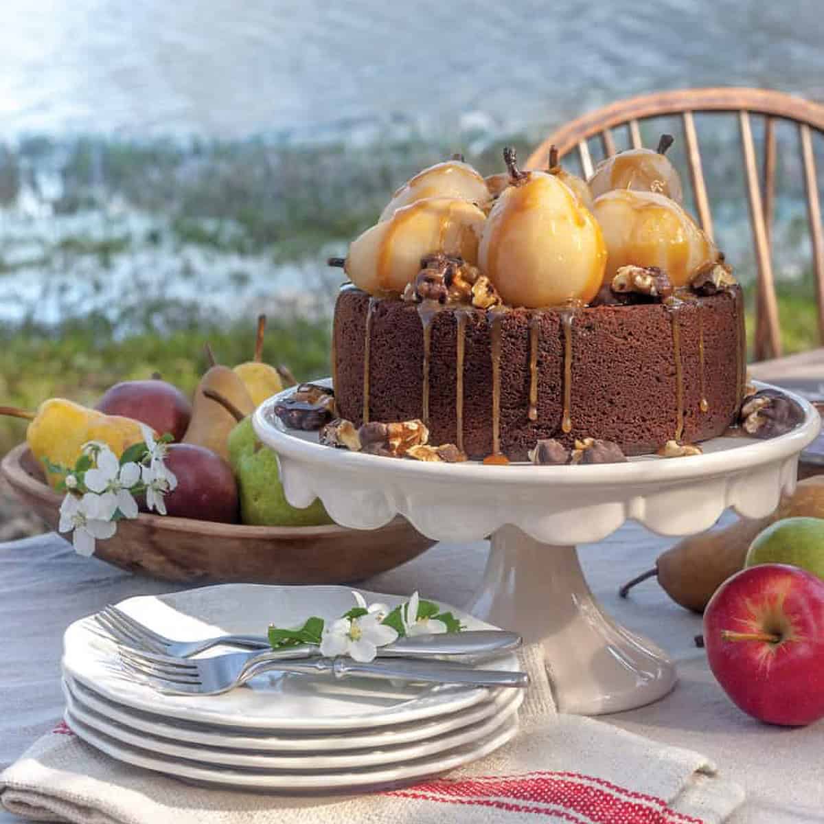 Chocolate cake topped with glazed pears and caramel drizzle on a white cake stand, surrounded by apples, pears, plates, and cutlery—perfect for an Autumn Afternoon Tea set outdoors by the water.