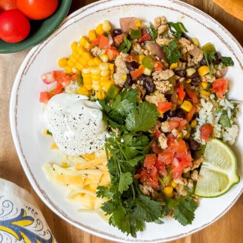 A plate with rice, ground meat, corn, beans, diced tomatoes, shredded cheese, sour cream, cilantro, and lime wedges-perfect for Taco Bowls. A bowl of tomatoes and tortilla chips are on the side.
