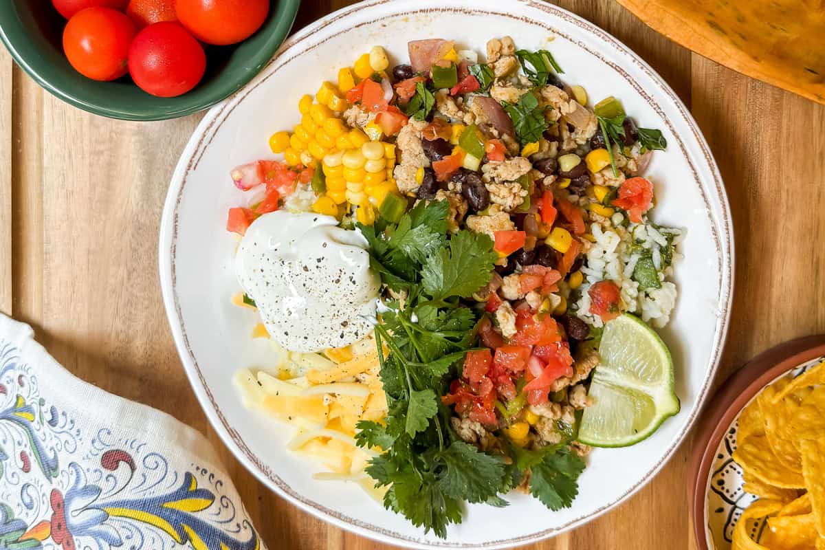 A plate with rice, ground meat, corn, beans, diced tomatoes, shredded cheese, sour cream, cilantro, and lime wedges-perfect for Taco Bowls. A bowl of tomatoes and tortilla chips are on the side.
