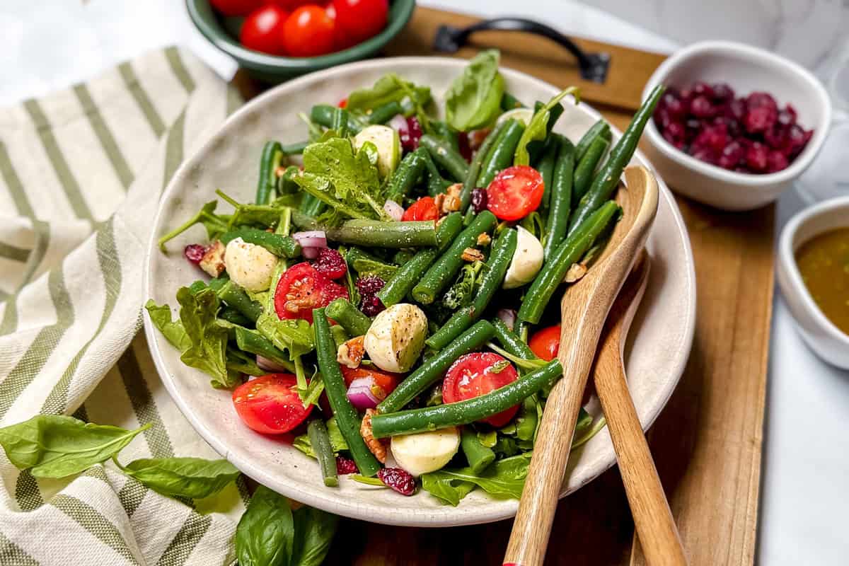 A vibrant Green Bean Salad with cherry tomatoes, mozzarella balls, greens, red onion, and dried cranberries is served in a bowl with wooden utensils.