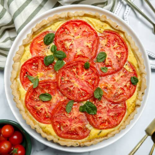 A baked tomato basil pie topped with sliced tomatoes and fresh basil leaves sits on a white plate with a striped cloth and cutlery beside it. Bowls of tomatoes and lemon wedges are nearby.