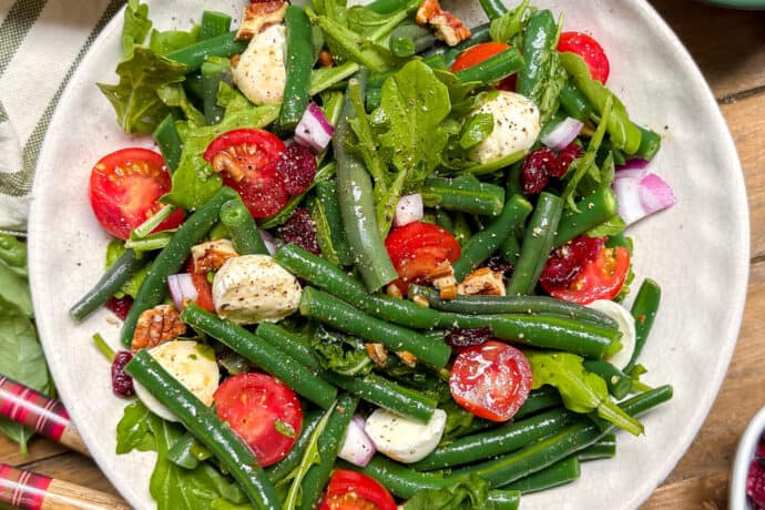 A vibrant Green Bean Salad featuring cherry tomatoes, mozzarella balls, red onion, arugula, pecans, and cranberries sits on a wooden table.
