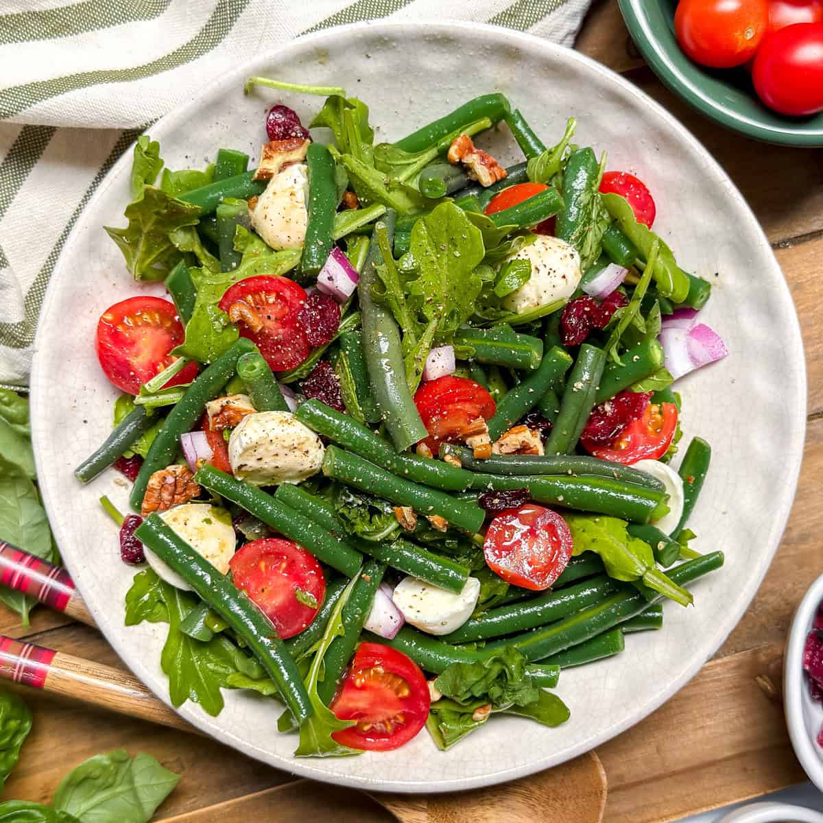 A vibrant Green Bean Salad featuring cherry tomatoes, mozzarella balls, red onion, arugula, pecans, and cranberries sits on a wooden table.