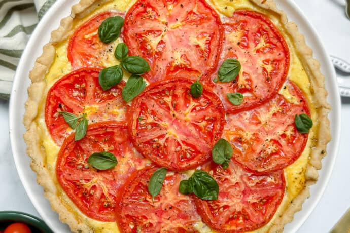 A baked tomato basil pie topped with fresh tomato slices and basil leaves, served on a white plate with a striped cloth and utensils nearby.