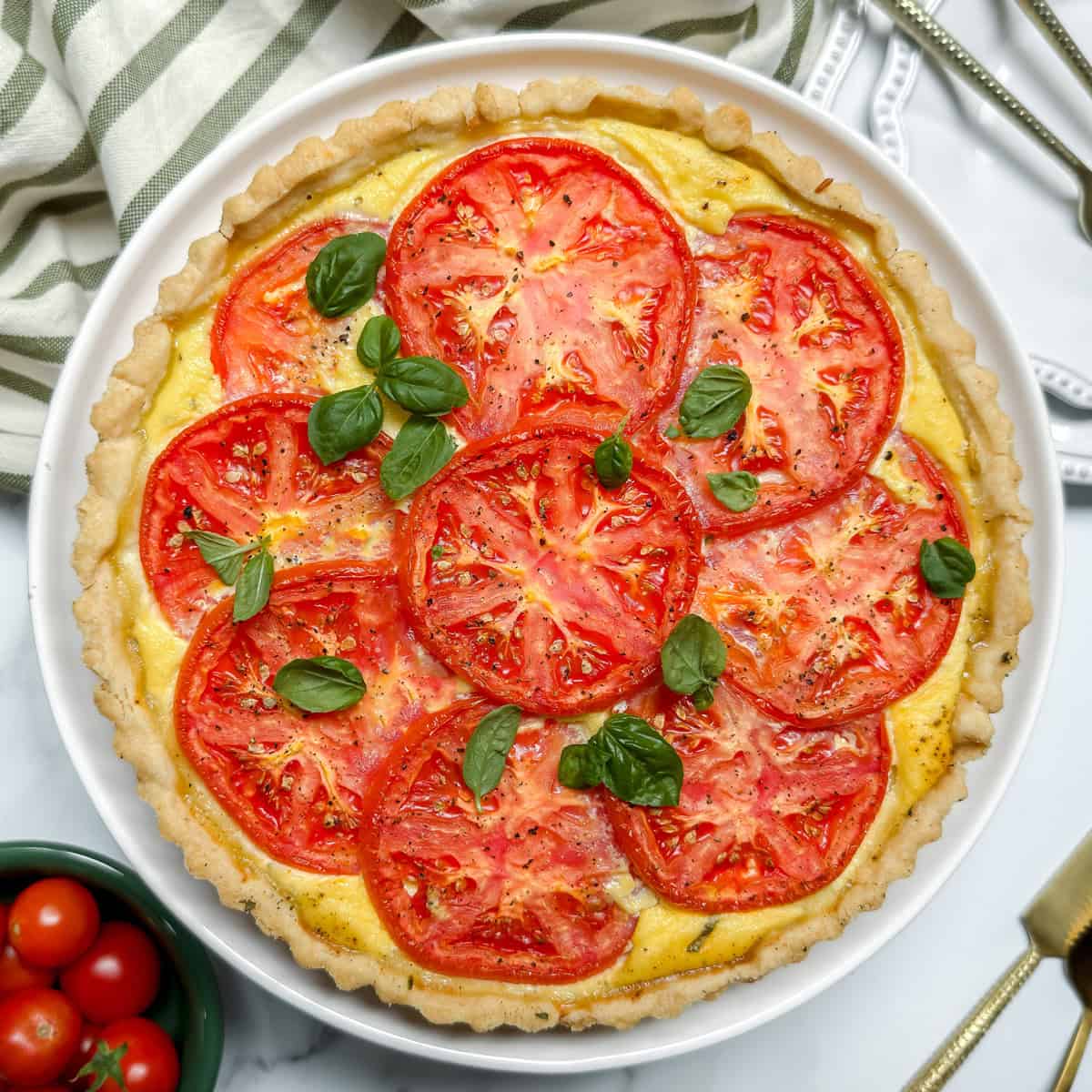 A baked tomato basil pie topped with fresh tomato slices and basil leaves, served on a white plate with a striped cloth and utensils nearby.