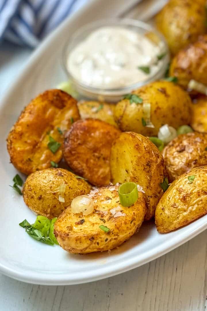 Close-up view of crispy air fryer potatoes with chopped green onions and herbs on a white plate with dipping sauce in the background.