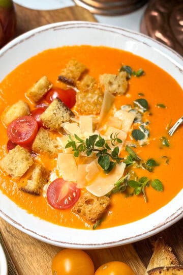 Creamy tomato soup in a bowl with a spoon and topped with Parmesan, croutons and fresh herbs.