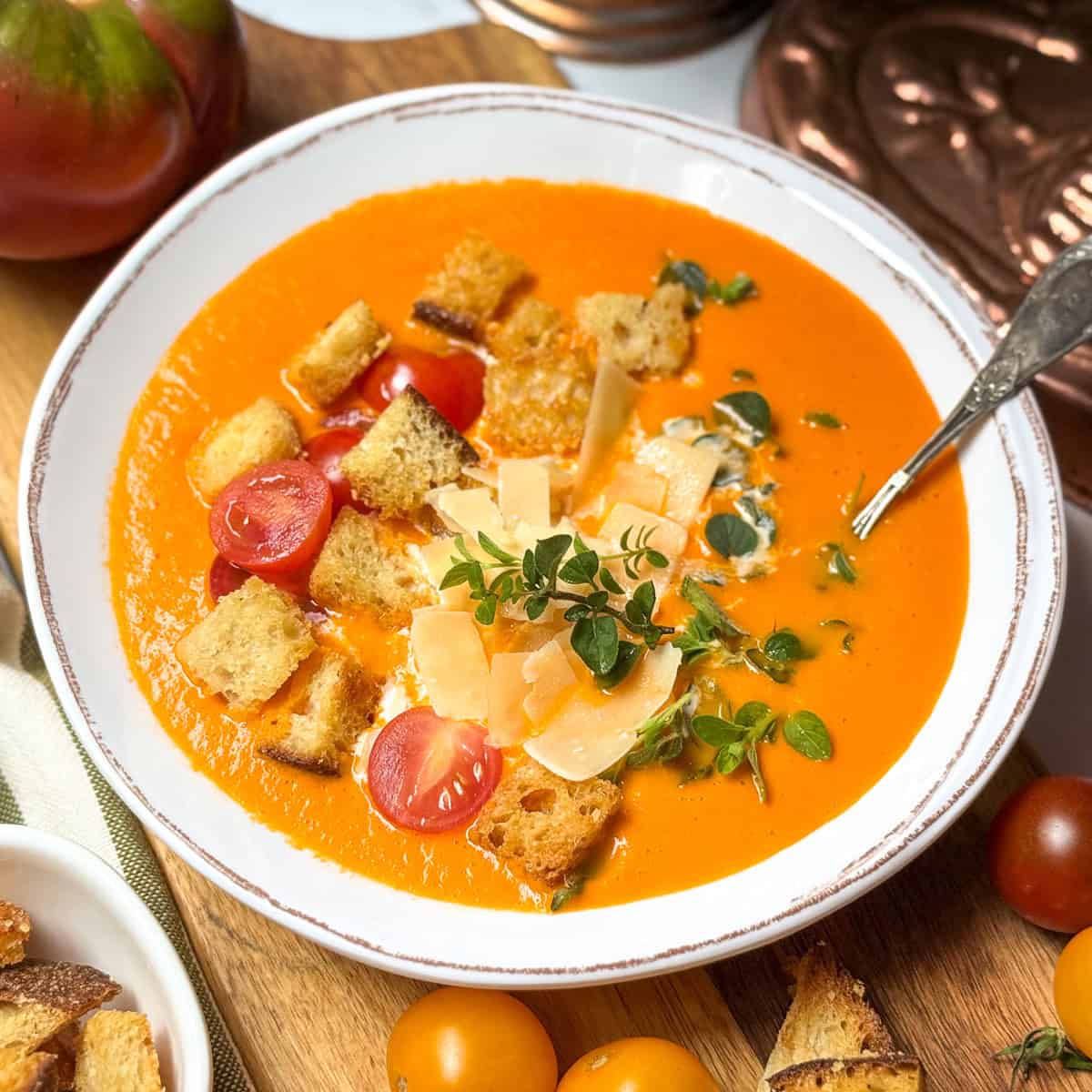 Creamy tomato soup in a bowl with a spoon and topped with Parmesan, croutons and fresh herbs.