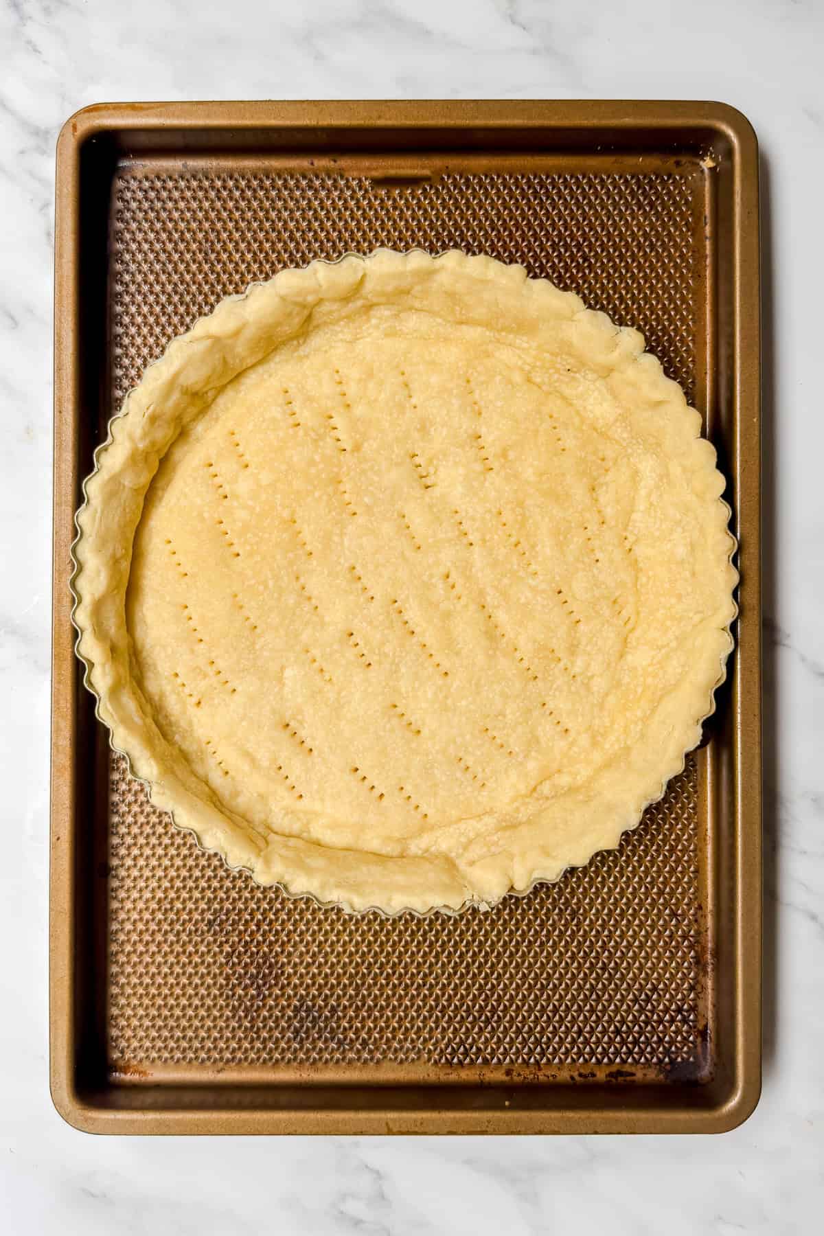 An unbaked pie crust in a tart pan sits on a baking sheet, with fork marks evenly pricked across the dough, ready to be filled for a savory tomato basil pie.