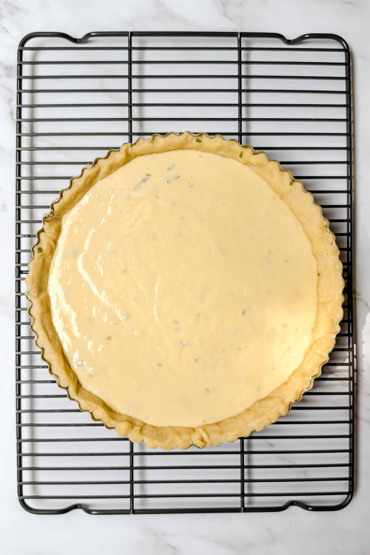 A tart crust for tomato basil pie filled with an uncooked creamy mixture sits on a black cooling rack atop a white marble surface.