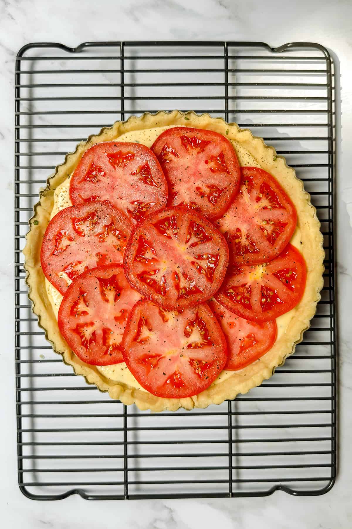 A tomato basil pie with fresh sliced tomatoes on a baked crust sits on a cooling rack over a marble surface.