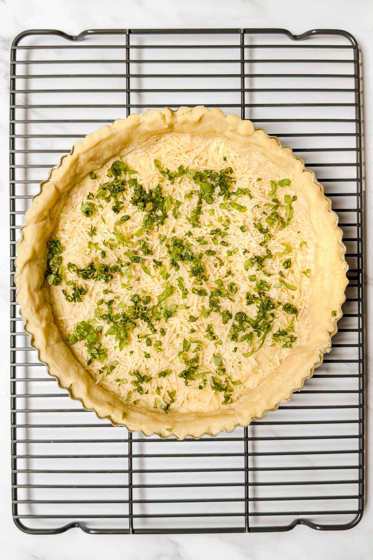 A partially baked pie crust in a tart pan, topped with shredded cheese and chopped herbs, sits on a cooling rack-perfect for a savory tomato basil pie.