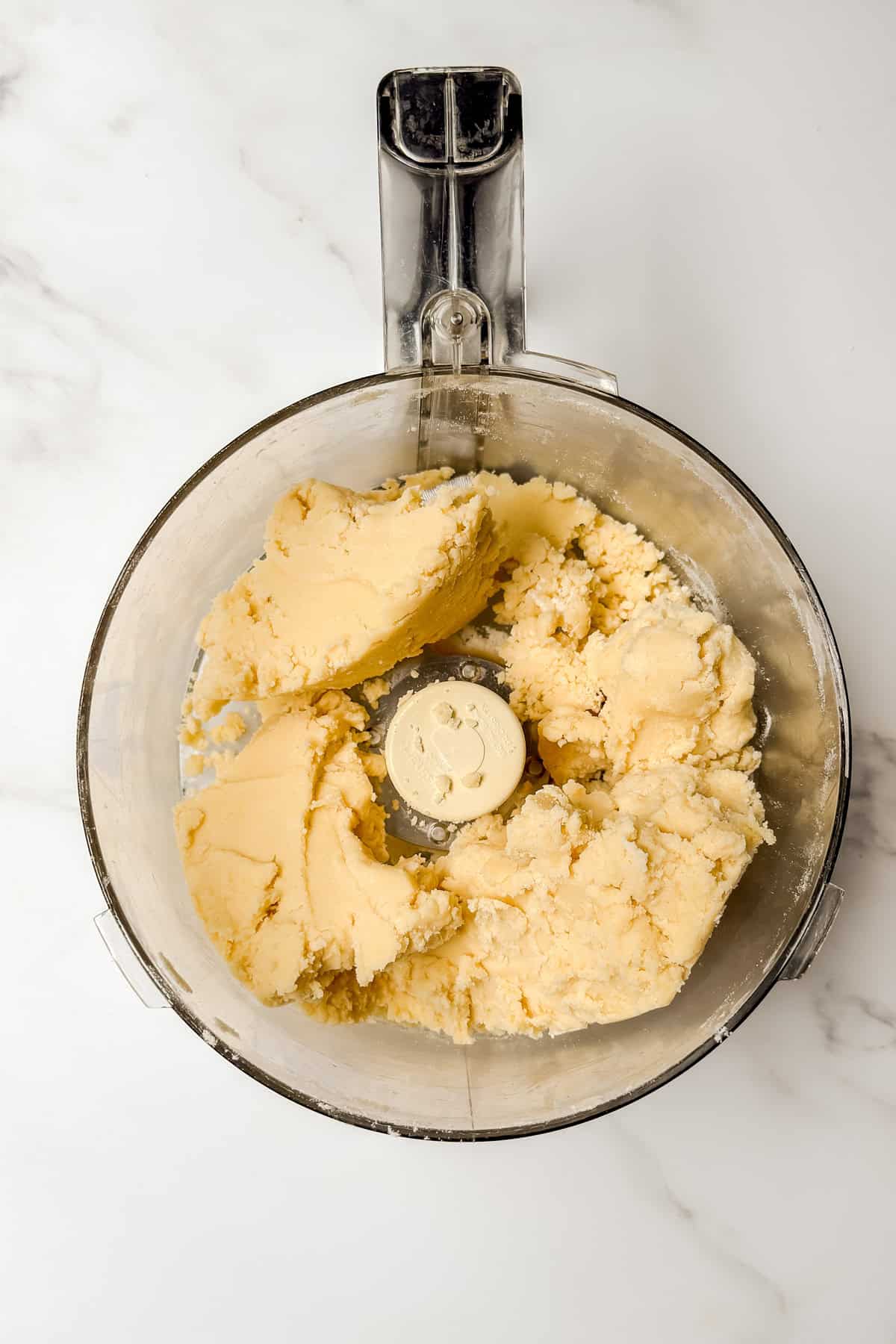 A food processor bowl containing pale yellow cookie dough, ready to be transformed into a delicious treat, sits on a white marble surface-perfect for pairing with a savory tomato basil pie.