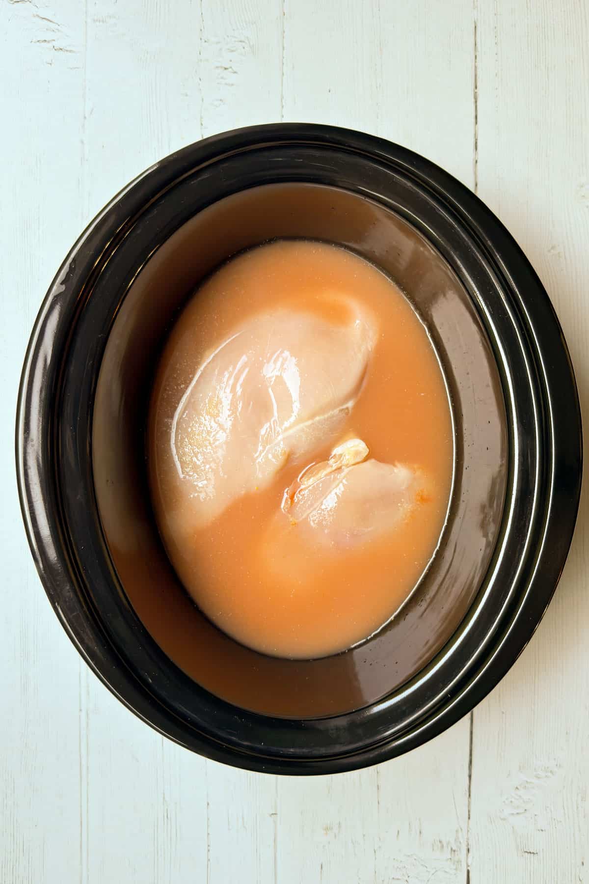 Raw chicken breasts with Buffalo sauce and broth in a black slow cooker.
