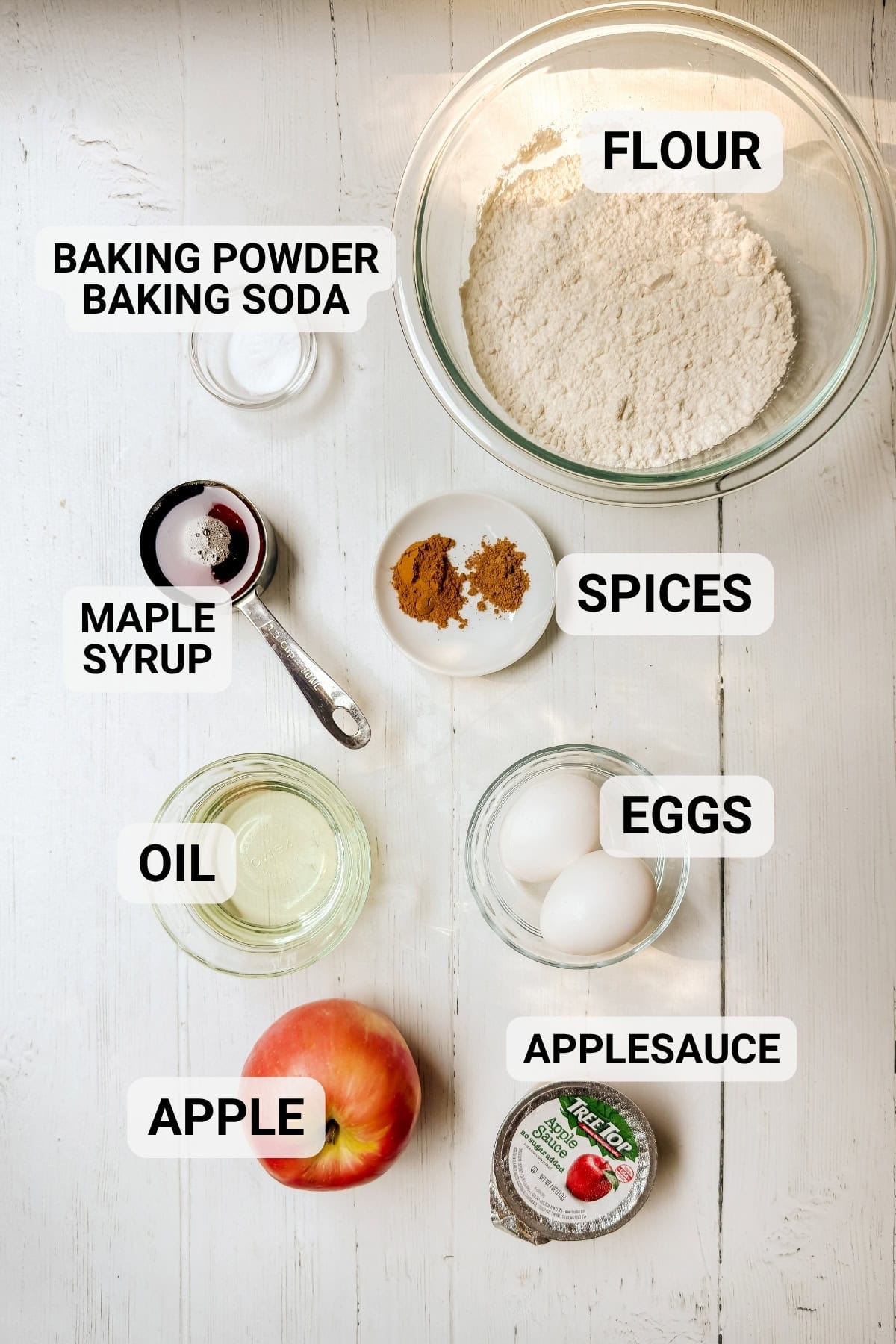 Ingredients for healthy apple muffins including flour, apple, applesauce, spices, maple syrup, and eggs.