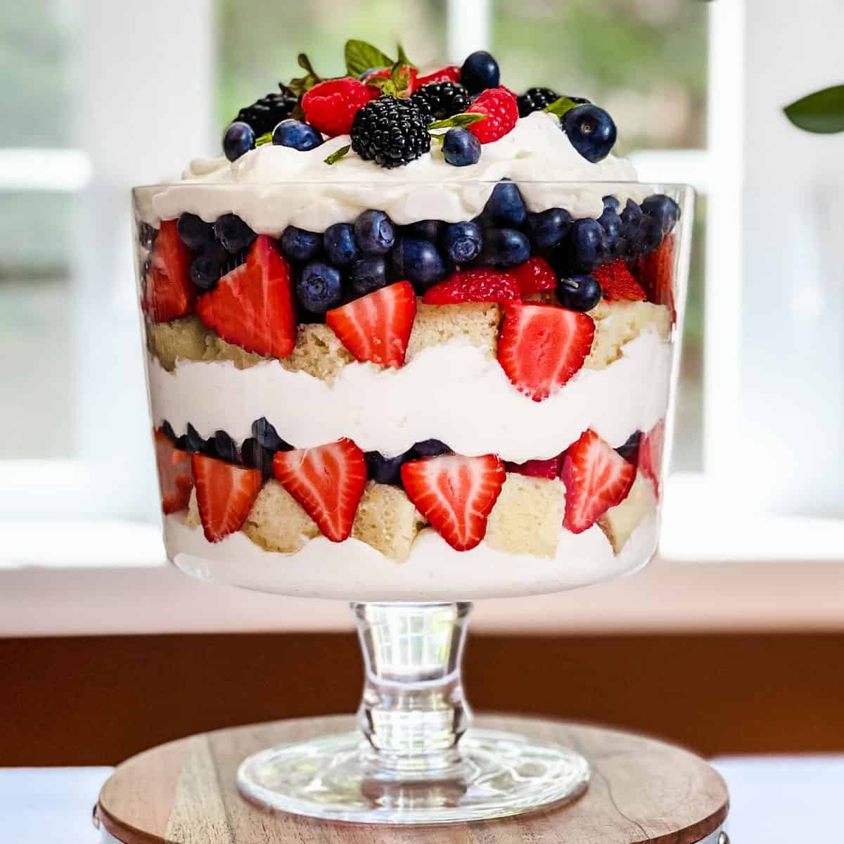 Square image of berry trifle with layers of fresh berries, chantilly cream, and fresh mint in a trifle dish ready for serving.