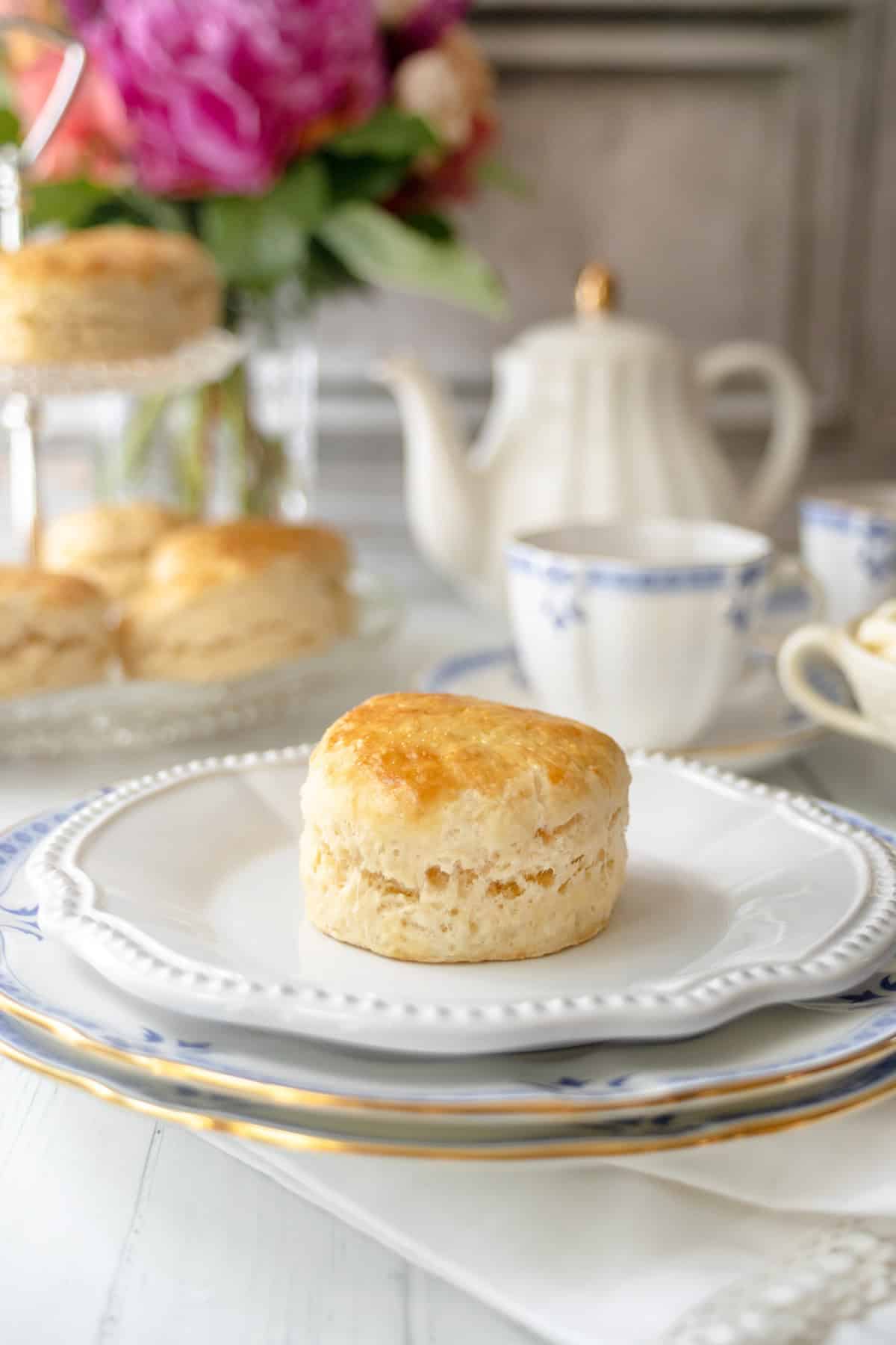 Golden baked scone on a plate set for afternoon tea with teapot and flowers.