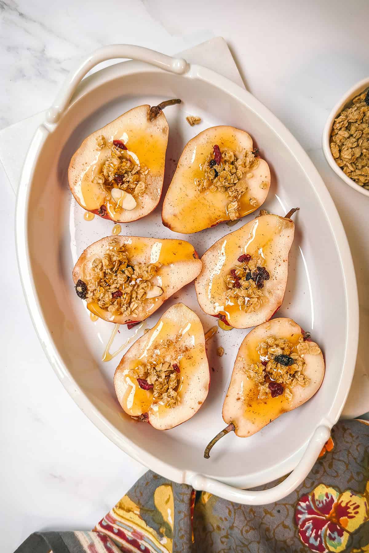 Halved pears, topped with granola and drizzled with honey in an oval baking dish ready for the oven.
