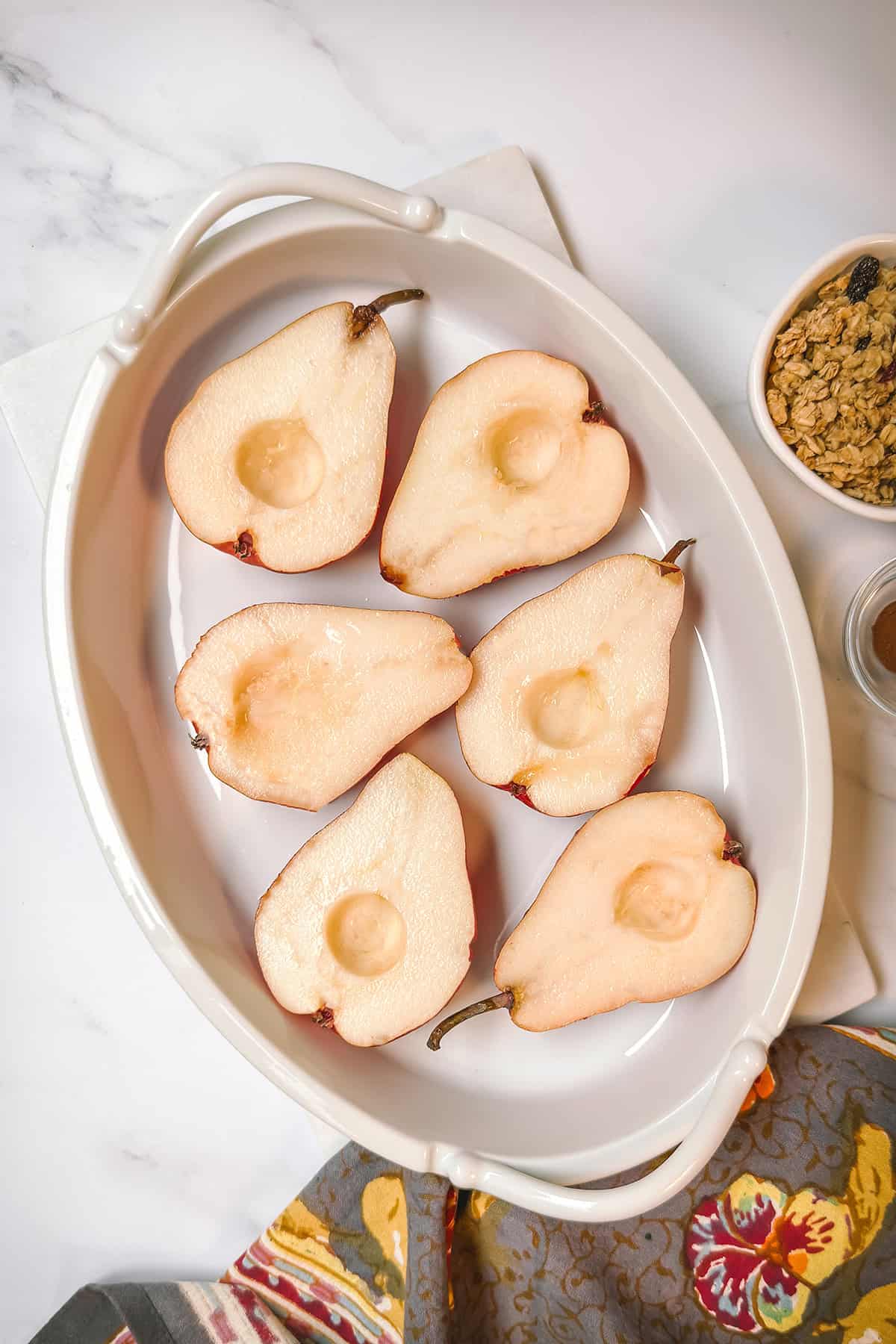 Halved and cored fresh pears in a white oval baking dish with granola and honey on the side.