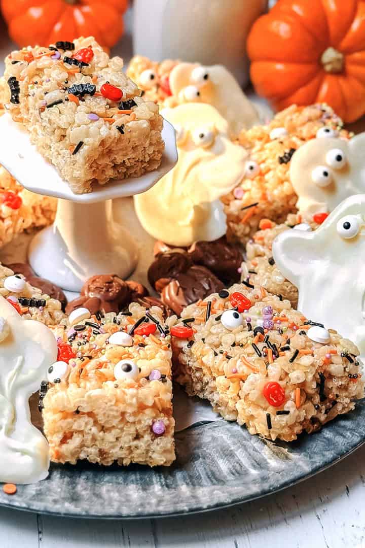 Squares of Halloween Rice Krispies Treats with white chocolate ghosts on a silver tray for serving with pumpkin in the background.