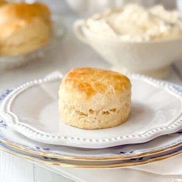 Golden baked English scone on a white plate with clotted cream in background.