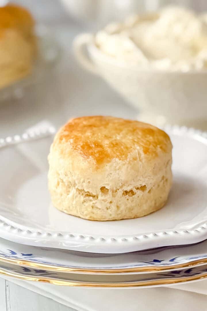 Golden baked English scone on a white plate with clotted cream in background.