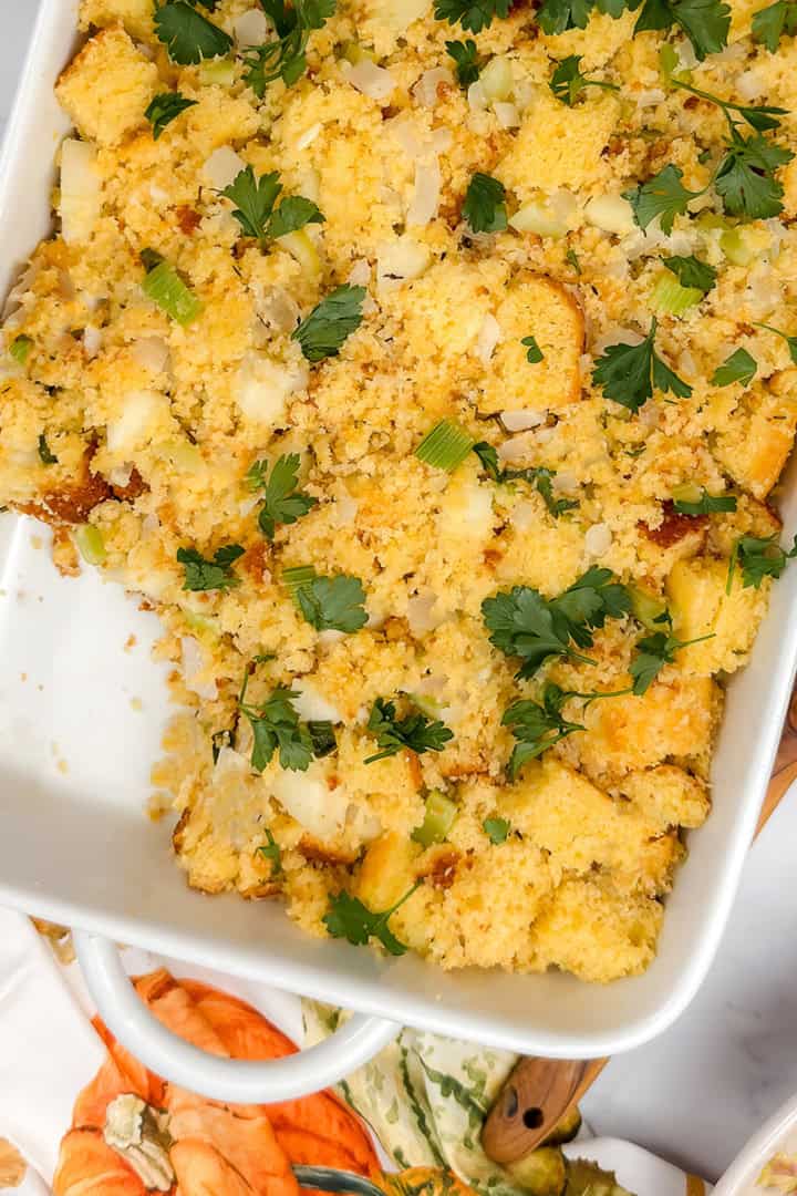 Square image of apple cornbread stuffing topped with fresh parsley in a white casserole dish with a portion missing.