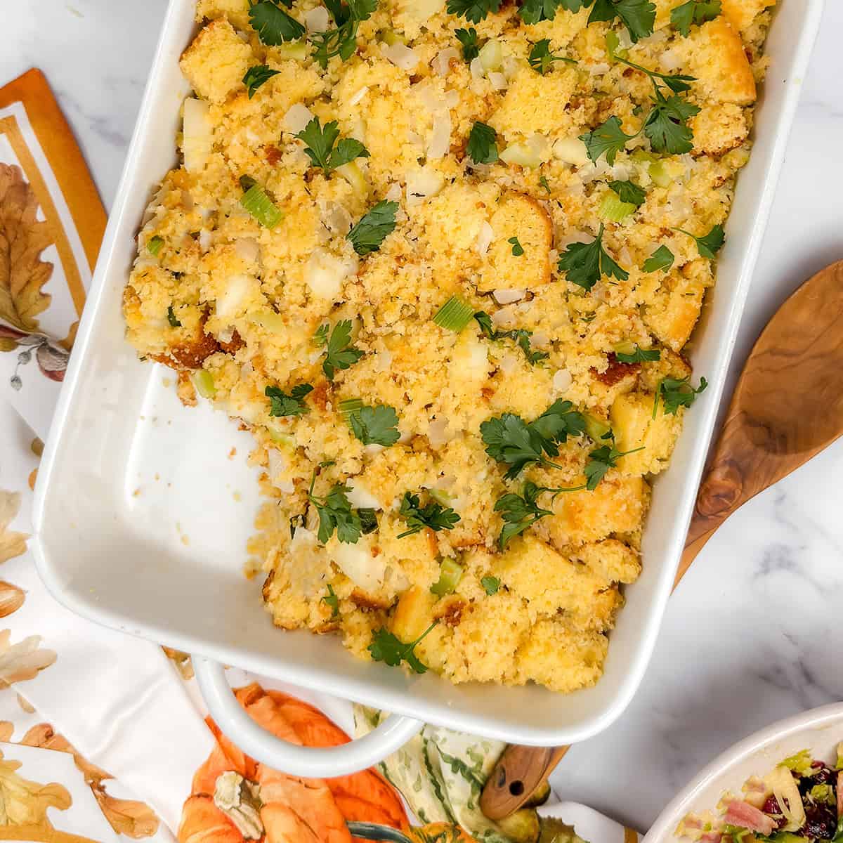 Square image of apple cornbread stuffing topped with fresh parsley in a white casserole dish with a portion missing.