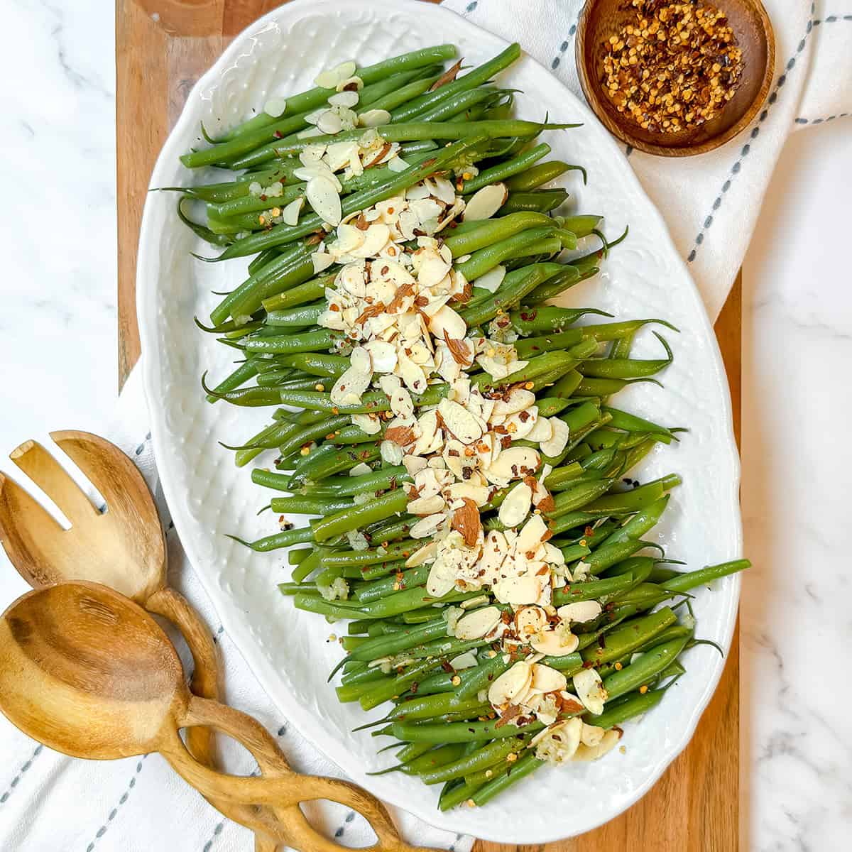 Square image of a platter filled with Green Beans Almondine arranged in rows and topped with toasted almonds and red pepper flakes.