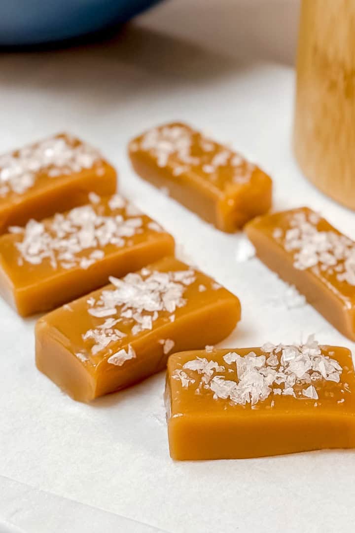 Square image of caramel candy squares topped with flaky sea salt on a parchment lined surface with wrapped candies is the background.